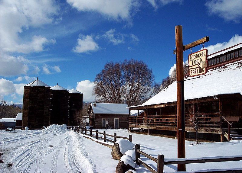 Snow-covered Pioneer Town sits to the right with a low fence. On the left in the distance three silos sit beneath a bright blue sky.