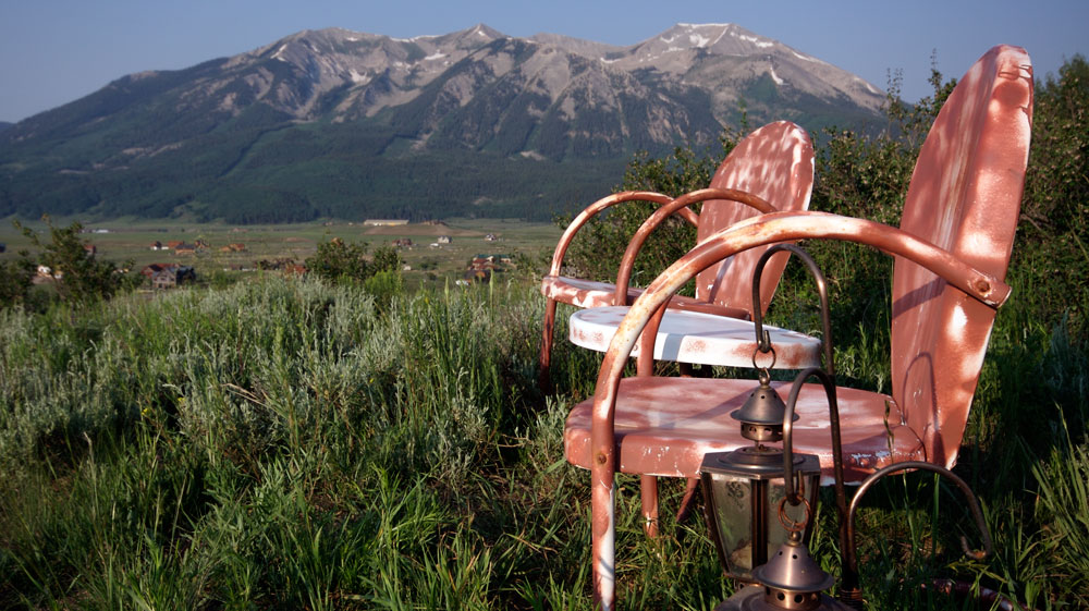 Two white but rusted-over metal lawn chairs sit in a field of tall, green grass. Between them is a small round, white metal table, also rusting but not as much as the chairs. In the background is a blue-and-gray mountain range underneath a dusty-blue sky.