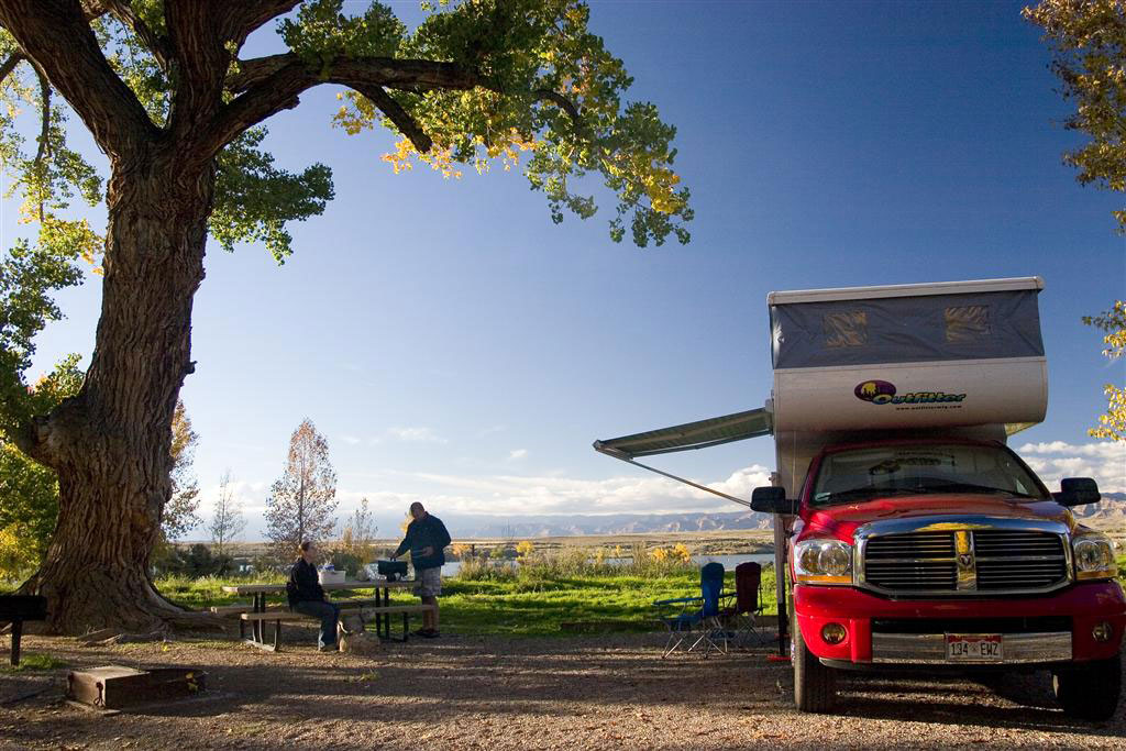 A dusty-blue summer sky glows with the warmth of the sun above a couple grilling something on a camp grill next to their red RV camper. They are in Colorado's Highlight State Park near Fruita.