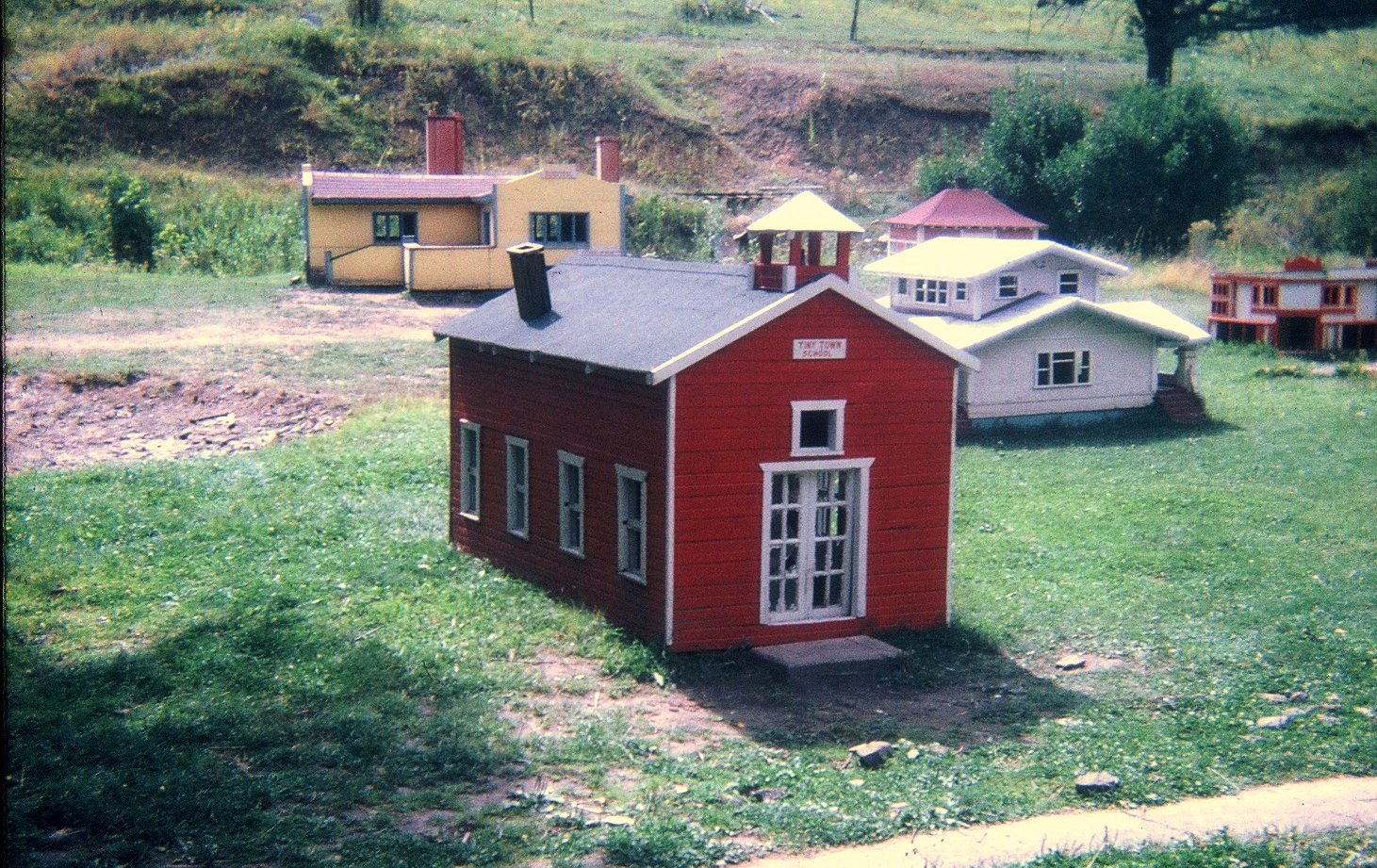 A miniature red schoolhouse sits on the grass among other small buildings in Tiny Town