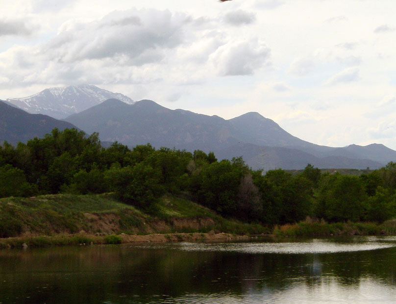 Fountain Creek Park's lake ripples on a summer's day surrounded by green grass and trees. A hazy, cloudy sky sits above Pike's Peak in the background.