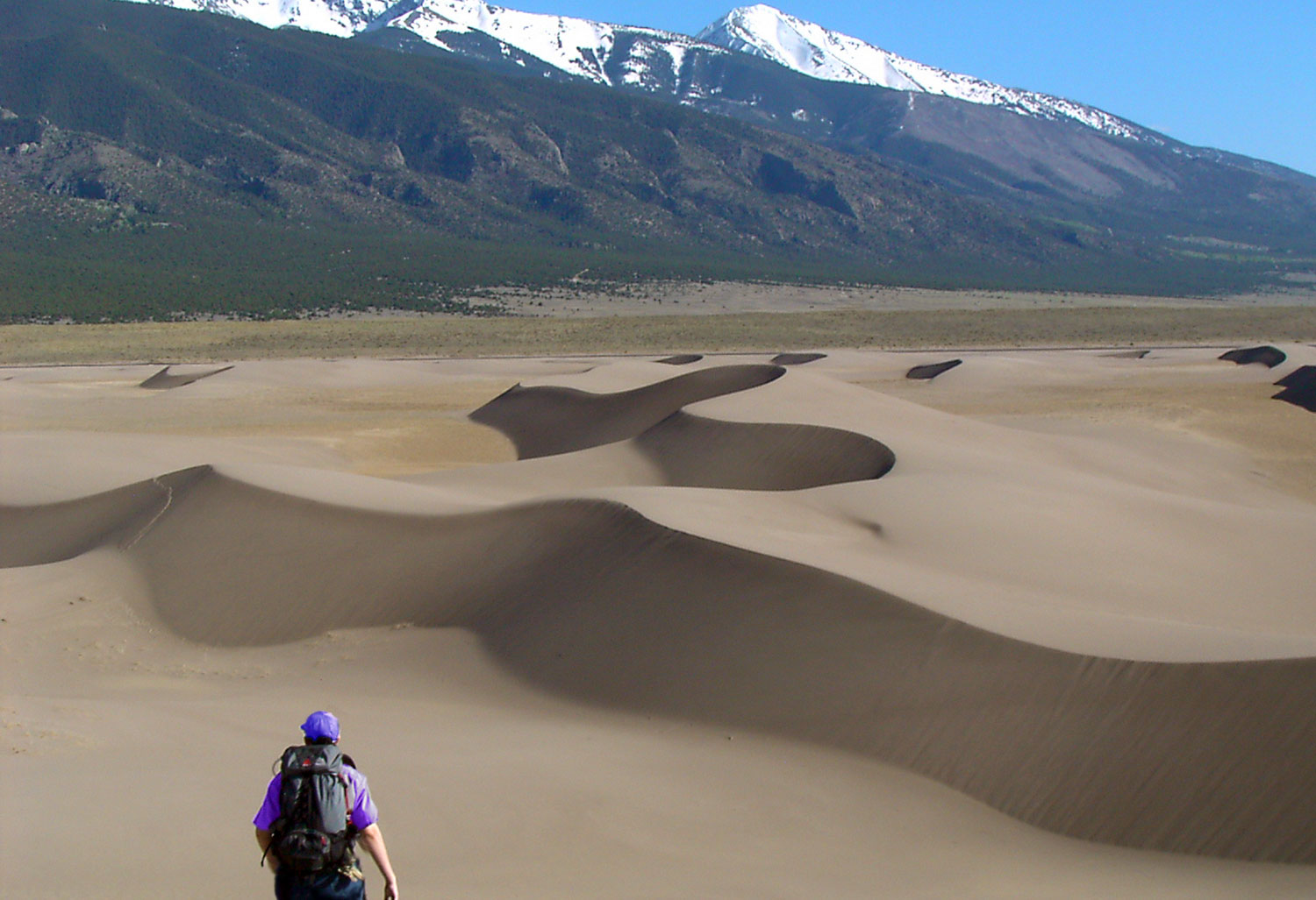 A person wearing a black backpack ventures out across the field of wavy sand dunes in Colorado. In the distance, mountain stand tall with peaks dipped in snow.