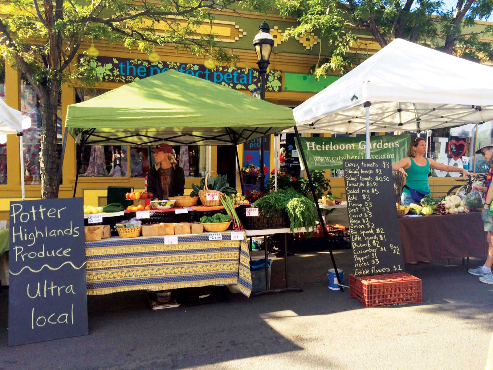 A chalk sign stands in front of two tented booths set up beside each other at the Vail Farmers' Market in Colorado. One is a vibrant lime-green and the other is white.