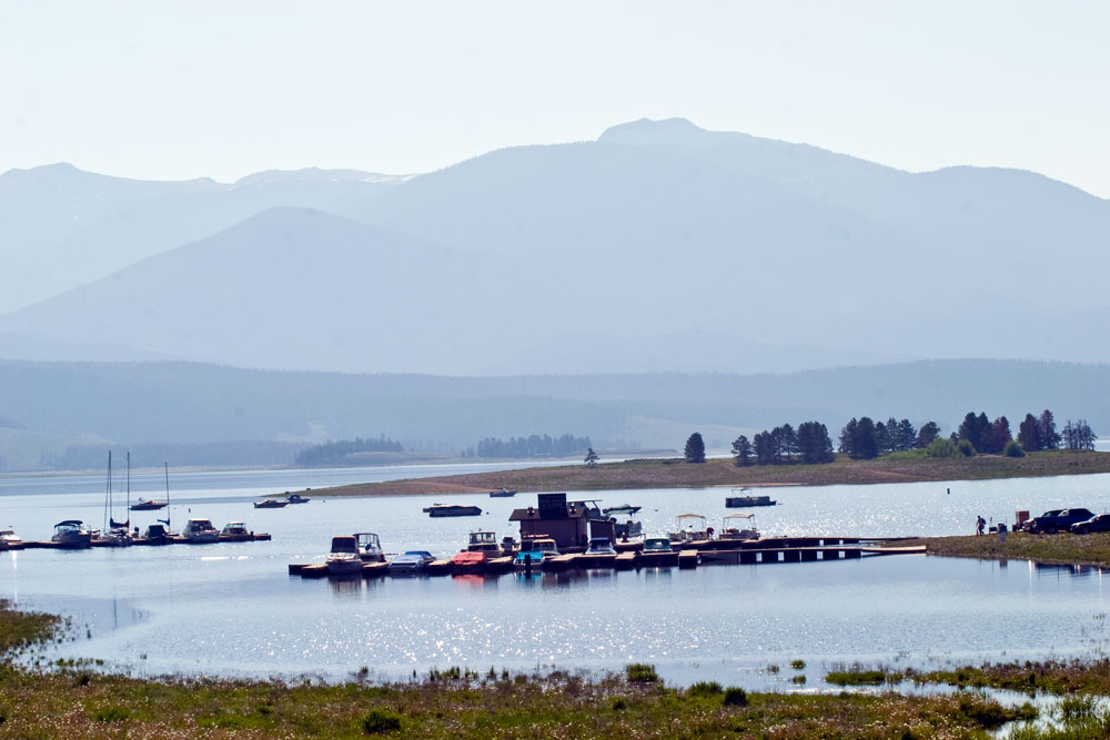 Boats are docked on a sparkling Lake Granby with hazy Rocky Mountains in the background.