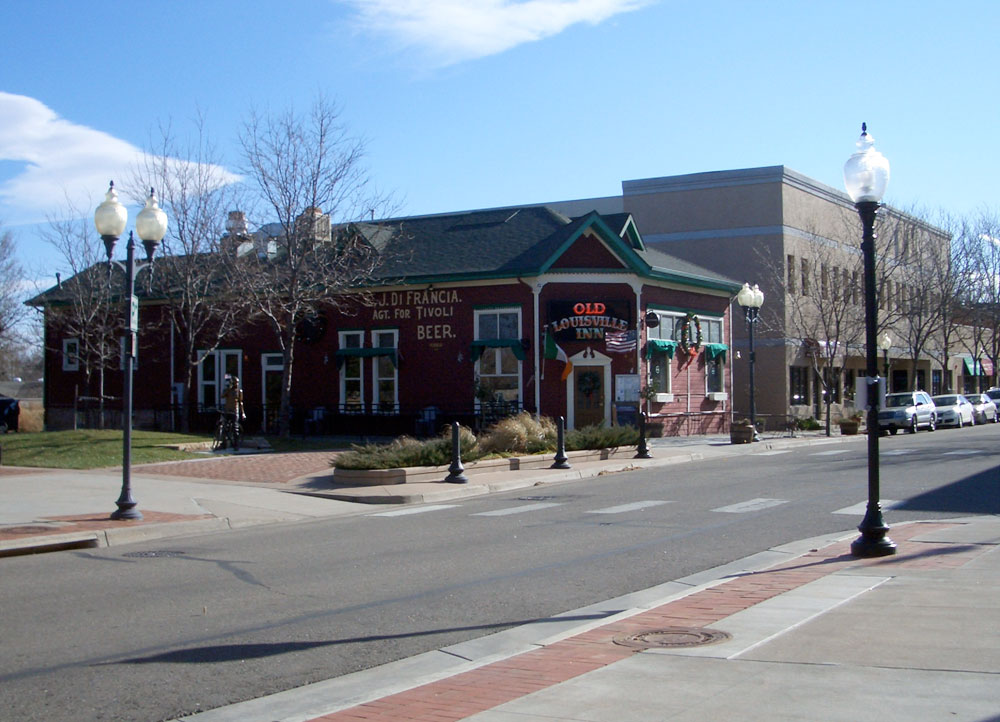 A view of the Old Louisville Inn from across the paved street. It's a red building with a green roof and a corner entrance. There's a two-story building next door and leafless trees line the sidewalk.