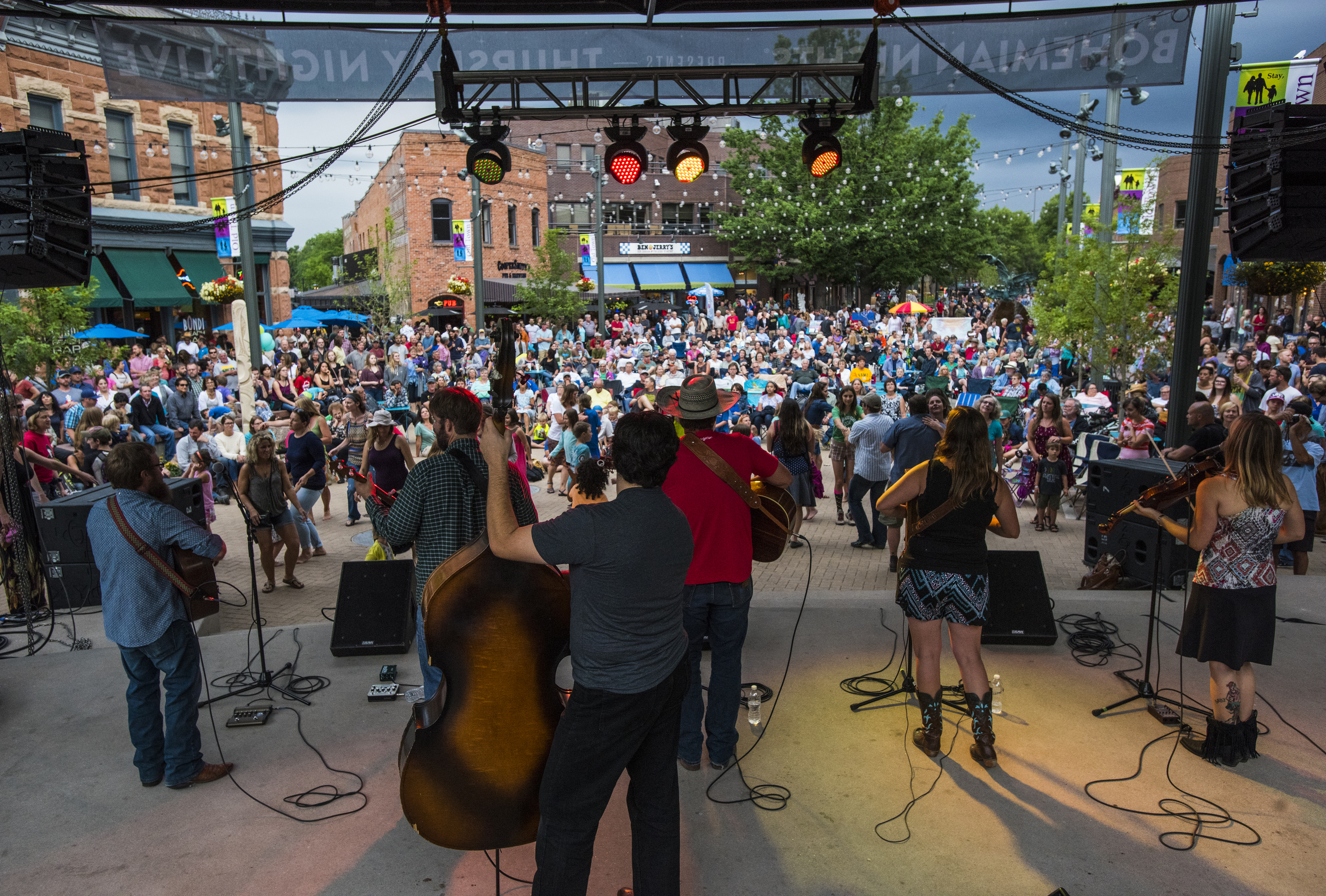A six-person band with their backs to the camera face a large crowd outside at Old Town Square. There are twinkle lights strung up between green trees.