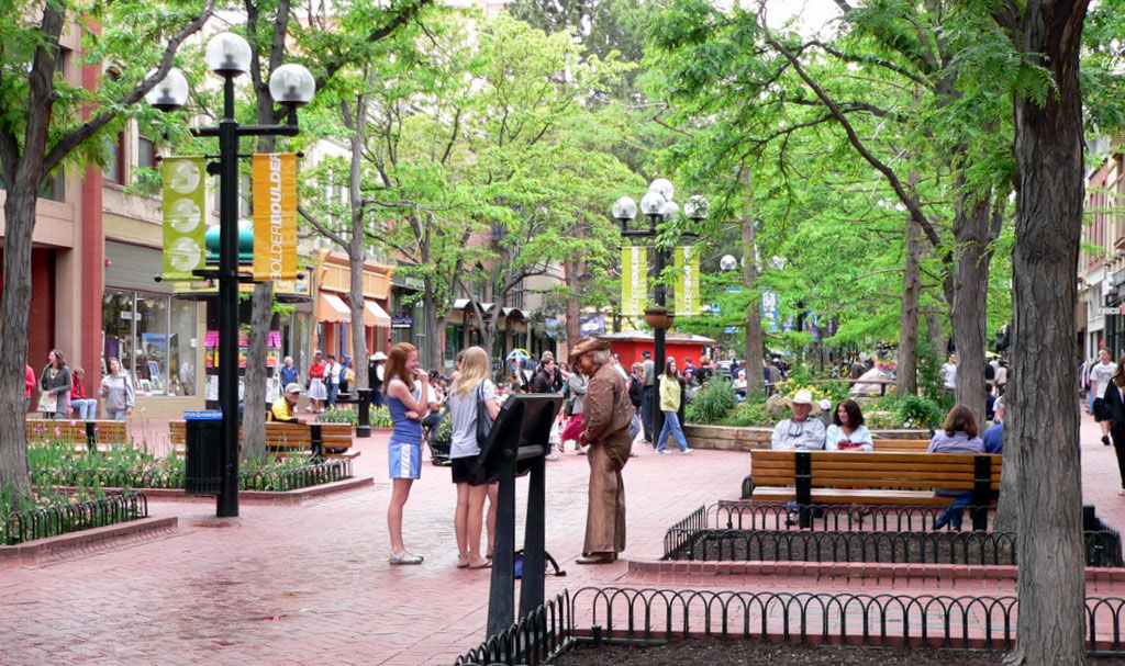 Green trees rise above the bricked pedestrian area of Boulder's Pearl Street Mall. The area features benches for sitting and many boutique shops.