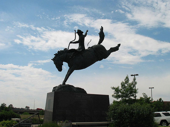 A life-size bronze sculpture of a bronco rider atop a horse stands in relief against a bright-blue sky peppered with white, fluffy clouds.