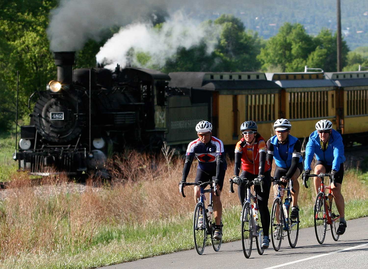 Four cyclists with helmets bicycle on a road alongside the Durango & Silverton Narrow Gauge Railroad under a bright summer sky.