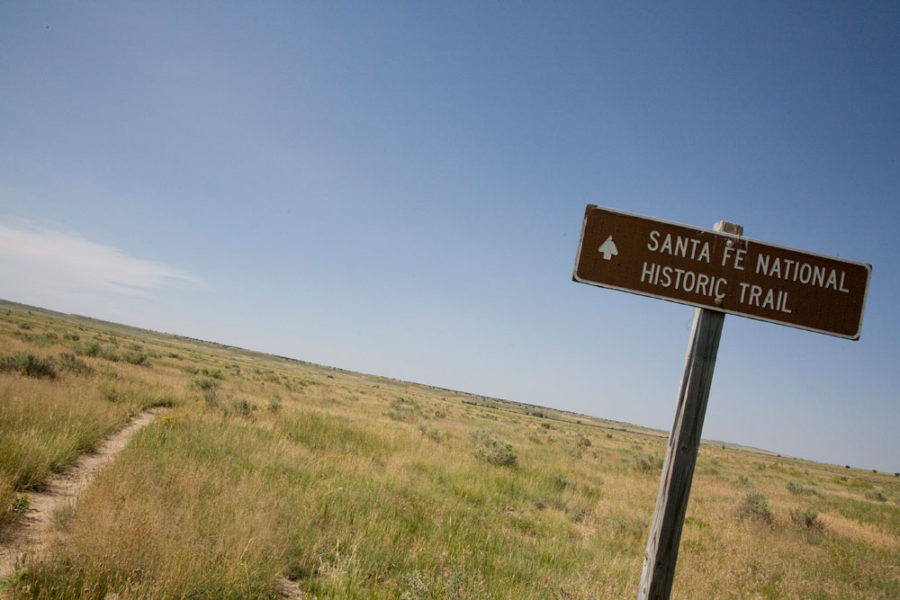 A tilted image of a metal brown rectangle sign that says "Santa Fe National Historic Trail" with an arrow pointing forward. There's a hazy blue sky in the background and the grown is covered in yellow and green tall grasses.