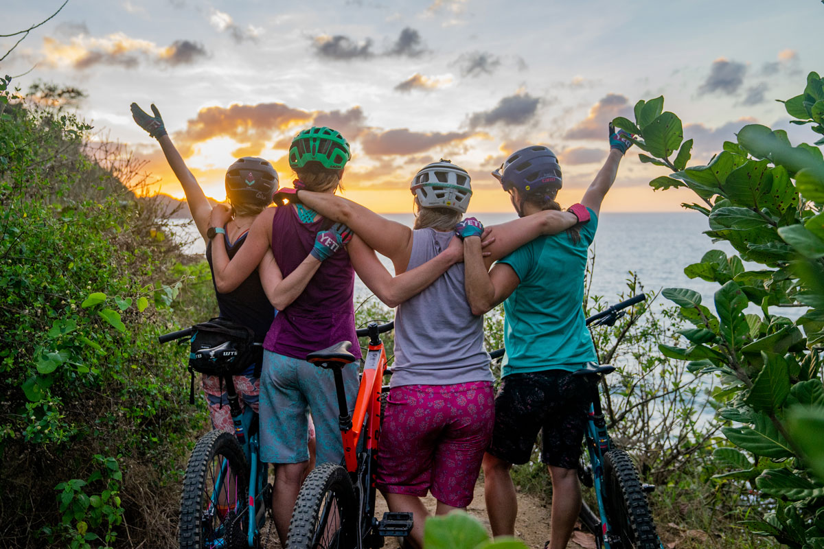 Four cyclists wearing outdoor gear drape their arms over each other or raise their arms to the run as they lookout over a lake in Colorado.