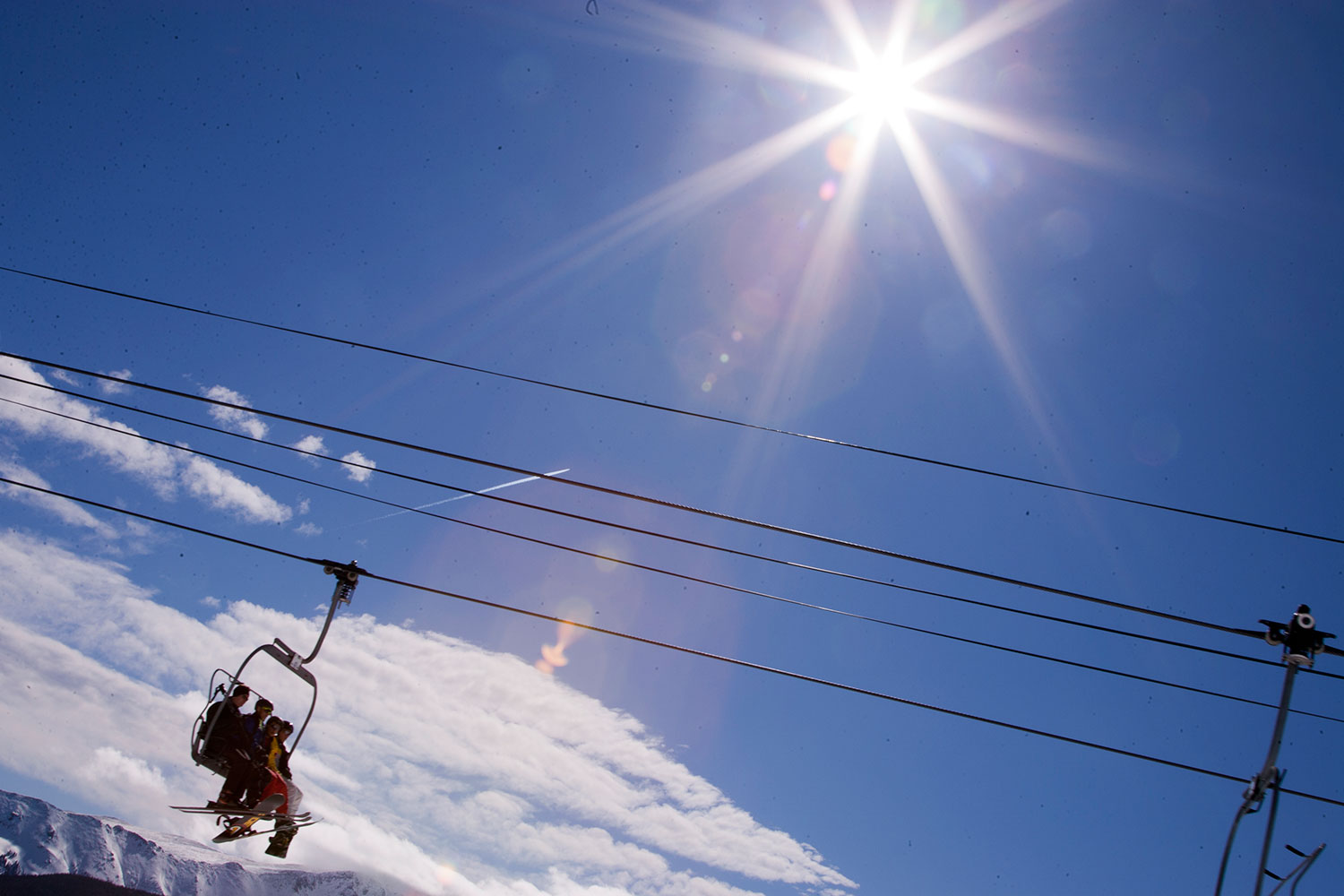 An angled shot of a chairlift with three people with skis on their feet moves through a blue sky with the bright sun above.