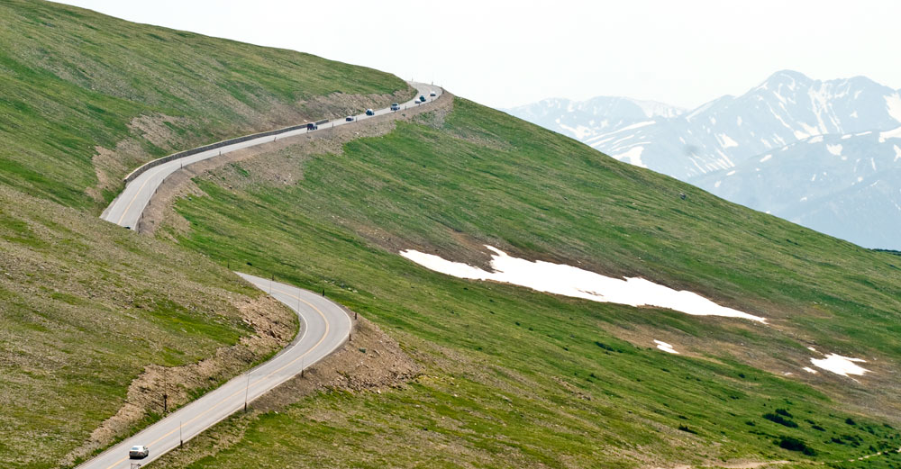 Cars drive along a two-lane highway wraps around the edges of a mountainside at Rocky Mountain National Park. There are patches of snow on the ground and on the distant mountains.