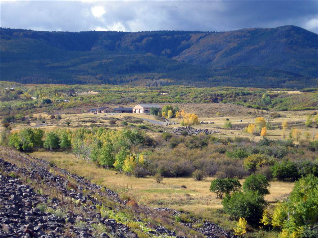 A long-distance view of the visitor's center at Vega State Park. In the background there are hills sitting under a stormy clouded sky. There are trees and scrubby bushes with tall grasses in the foreground. 
