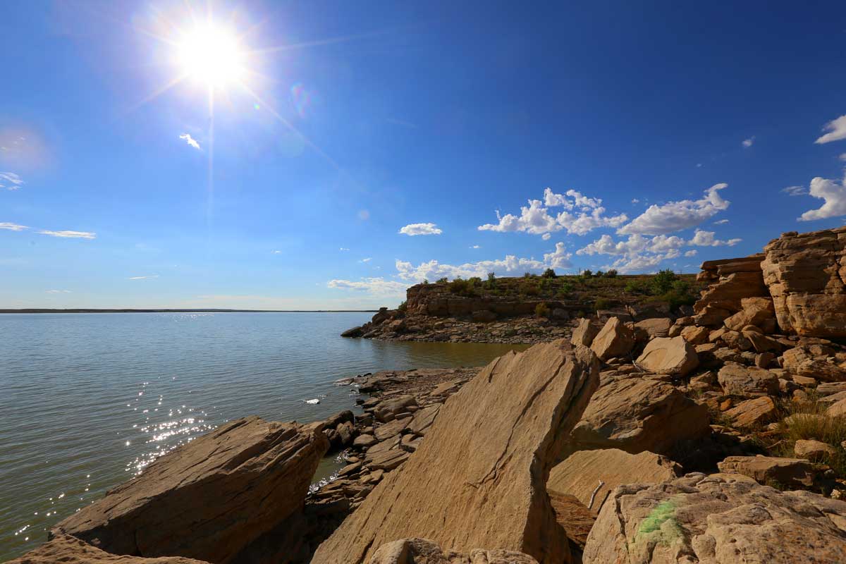The rocky shores of John Martin Reservoir are bathed in the bright sunshine. The sky is blue with a few clouds dotting the horizon and the water is rippling.