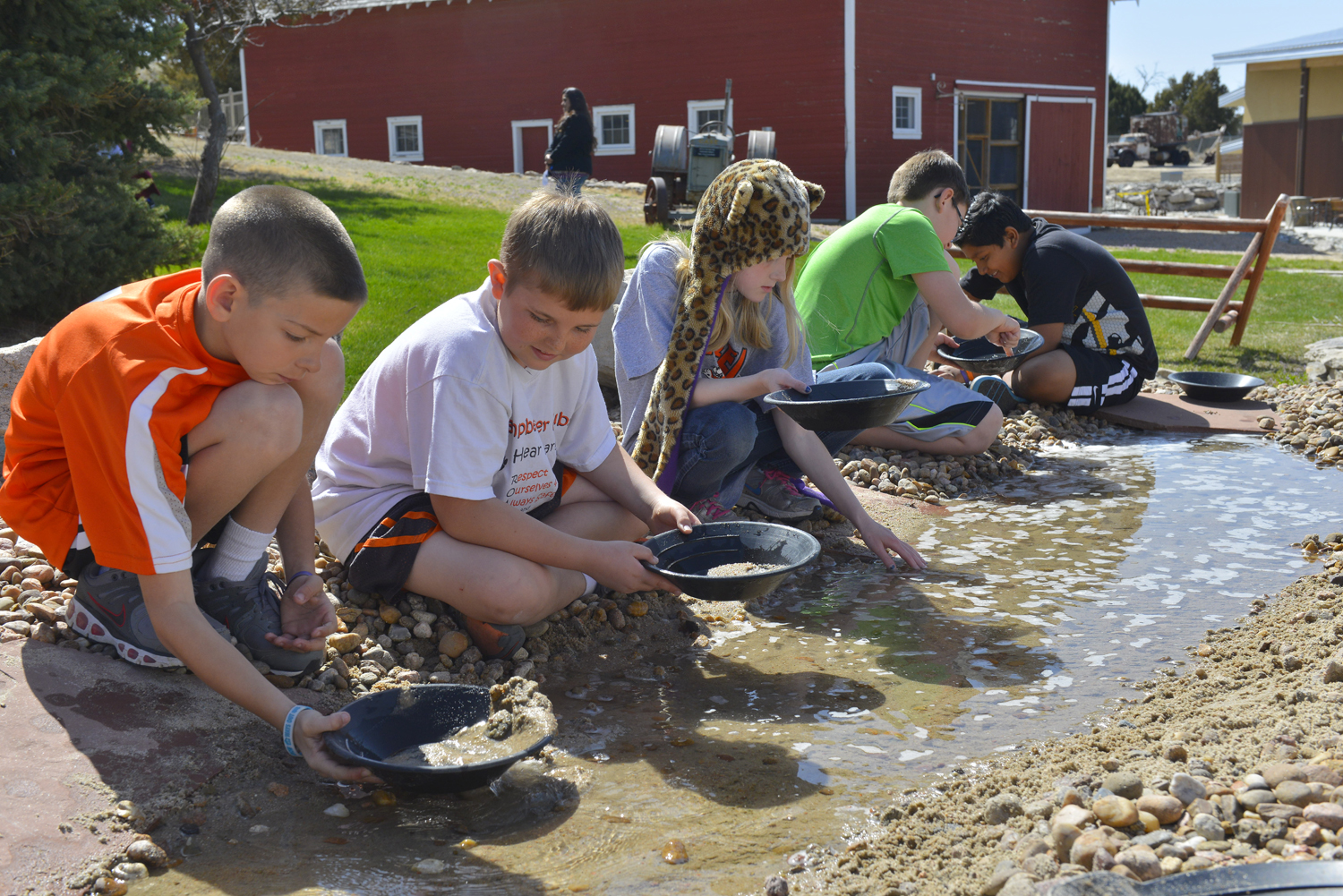 Five children pan for golden in a sudsy stream in front of a red building.