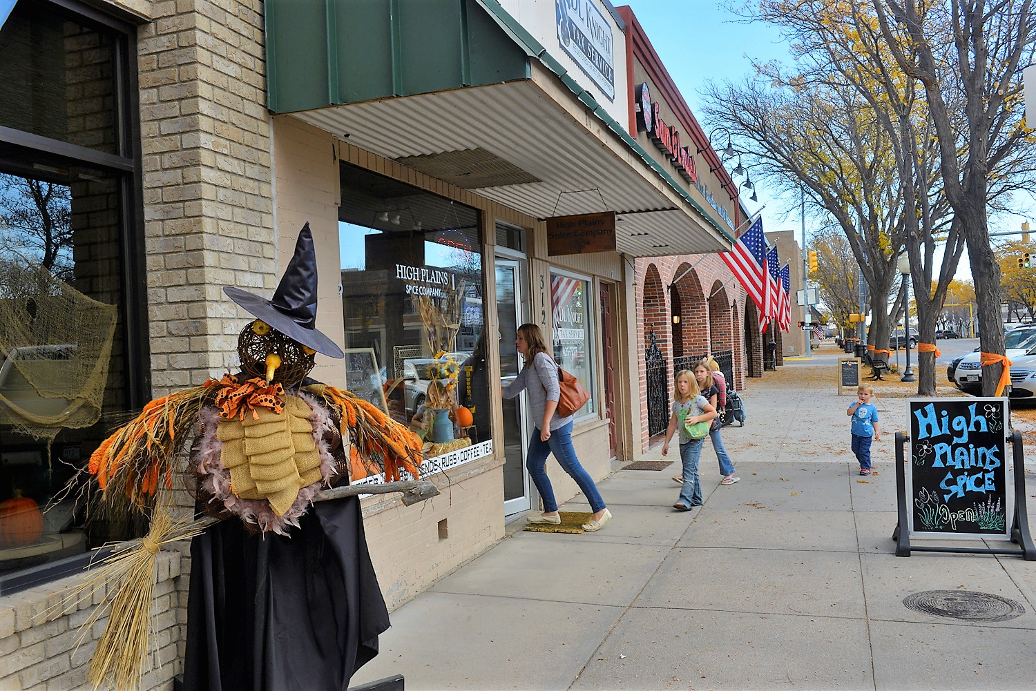 A family walks along a sidewalk with the mother entering a shop in downtown Sterling. On the right is a chalkboard sign that reads "High Plains Spice." On the left is a scarecrow dressed as a with with a bird's face.