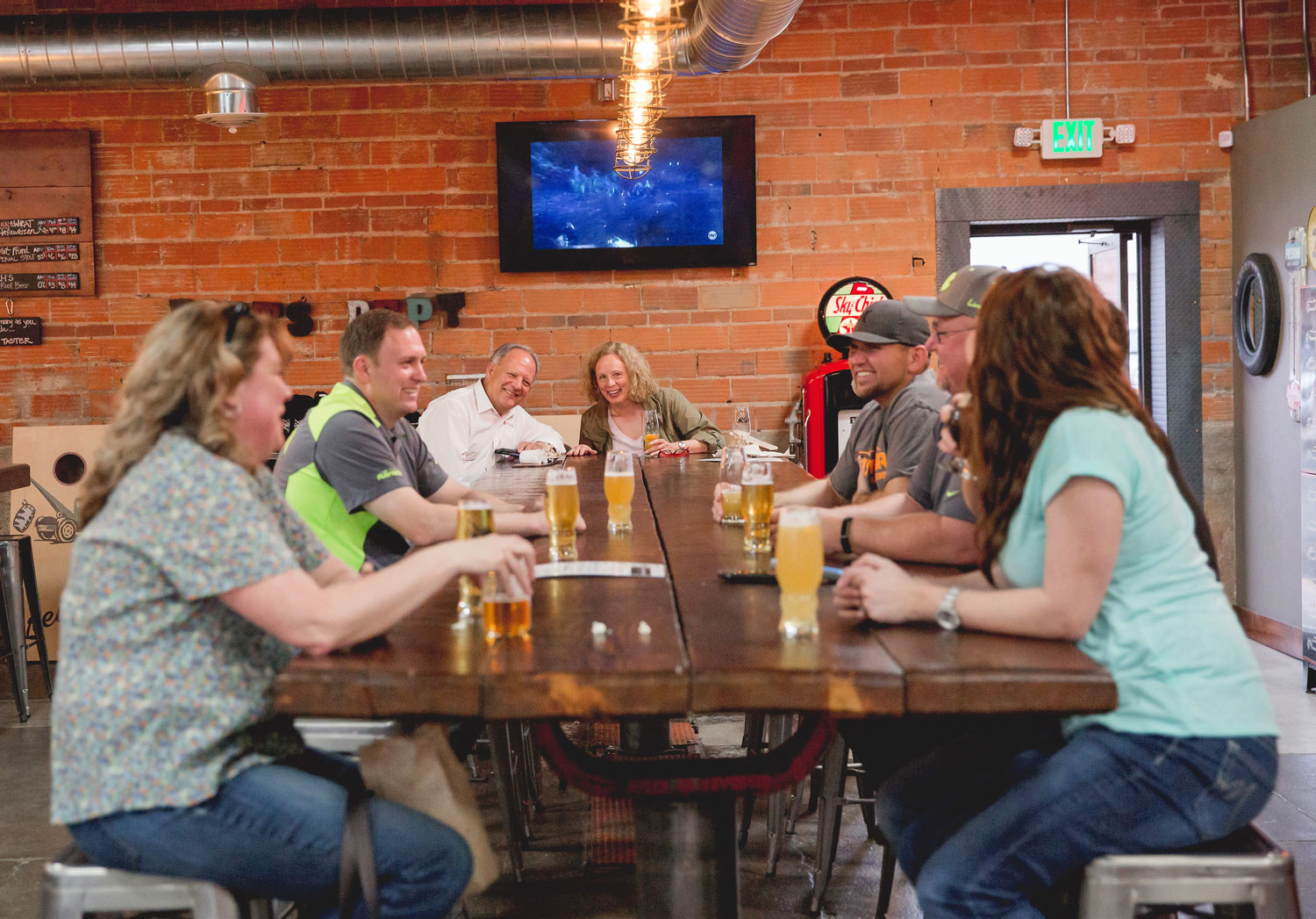 A large group of people sit at a wooden table as they drink craft beer. There's a red-brick wall with a TV in the background.