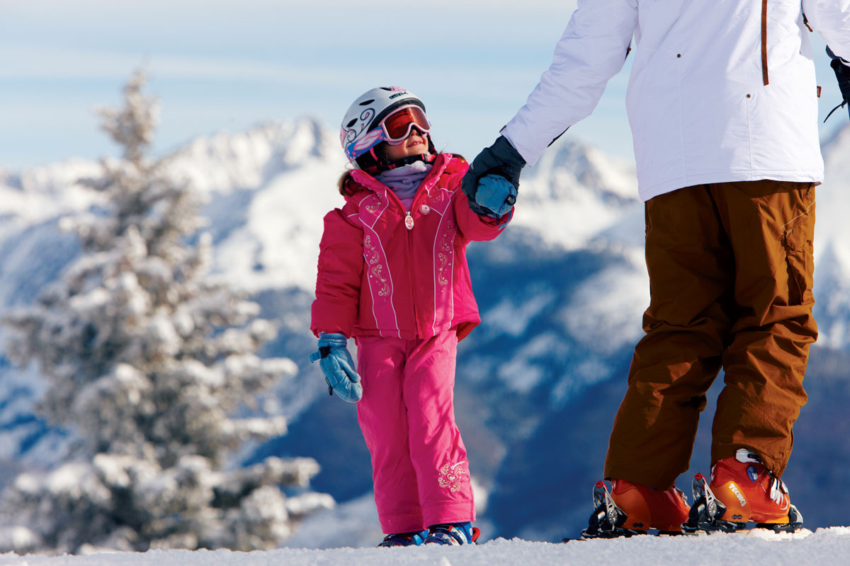 A small child smiles while holding an adult's hand at Vail Ski & Snowboard School on Vail Mountain with snowy peaks in the background
