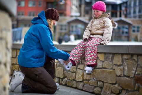 A young child sits on a stone wall as an adult helps them put on white ice skates at Winter Park Resort.