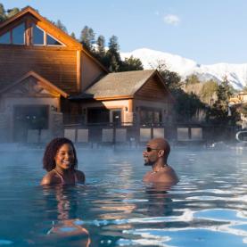 Couple soaks in a hot-springs pool as steam rises off the water. Snow-covered mountains are seen in the distance.