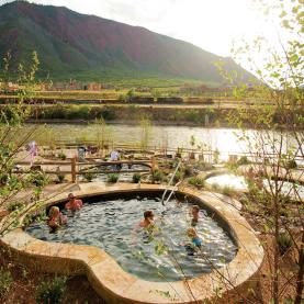 A group of people soaks in a stone hot pool overlooking a river and mountains