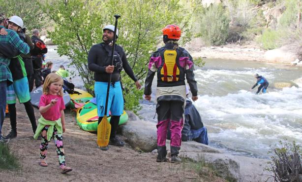 Paddlers standing on the banks of the Arkansas while a person surfs a rapid