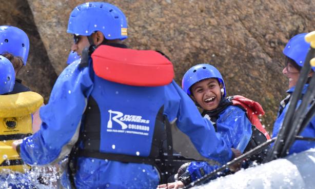 A photograph of a kid smiling widely, clad in a blue helmet, blue jacket and a life vest. They are on a whitewater rafting trip with their family on the Arkansas River in Colorado. Beside the kid are several smiling adults. There is water splashing up as they raft down the river.