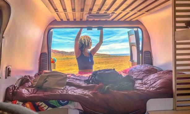 Woman closes door on the back of a campervan with cozy bedding and there are prairie grasses and blue skies in the background