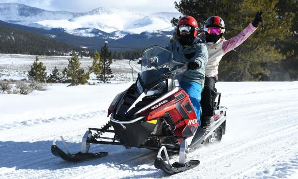 Two people wearing helmets, goggles and winter gear ride a red snowmobile near Leadville with snow-covered mountains in the distance.