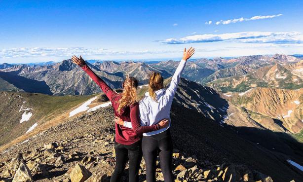 Two women at the top of a hike facing mountains with a blue sky above.