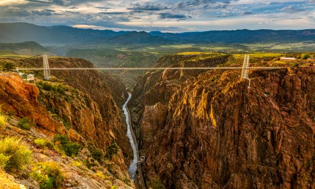 Royal Gorge Bridge & Park: Golden hour with Arkansas River snaking through rock walls of the gorge with the metal suspension bridge across the upper part of the canyon.