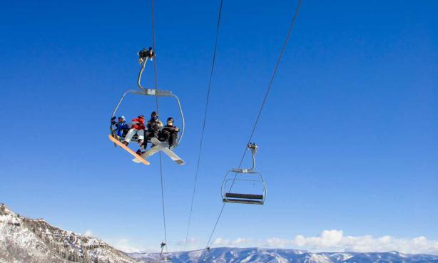 Snowboarders and skiers ride a chairlift at Snowmass Mountain with a vast bluebird blue sky behind them