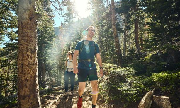 Two hikers on a trail, surrounded by trees, hike along in the sunlight