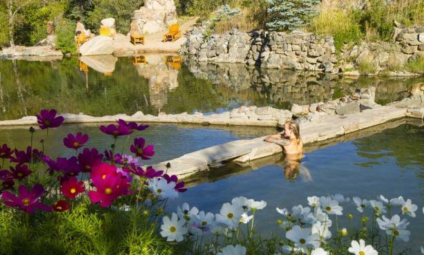 A woman rests on the edge of the Strawberry Park Hot Springs pool on a sunny day, with pink and white wildflowers blooming in the foreground