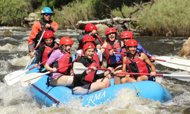 A group of red vest- and helmet-clad younger people sit in an inflated blue raft on the rolling whitewater waves of the Cache la Poudre River in Colorado. They are smiling and holding paddles in their hands while the instructor in the back of the raft guides them along the water. In the background are green plants and trees lining the river.