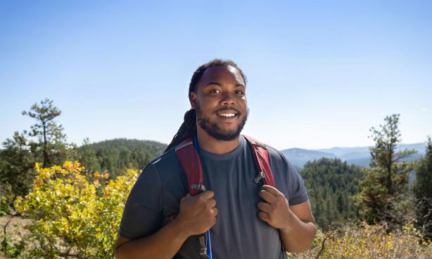 Colorado hiking enthusiast Nelson Holland stands holding his backpack straps in front of a mountainous backdrop.