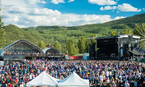 A crowd gathers at two stages with green mountains behind them to listen to bands at the JAS Labor Day Experience in Snowmass