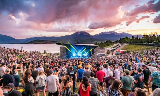 A crowd listens to a pand perform from a blue-lit stage with Dillon Reservior, mountain peaks and pink clouds as the backdrop