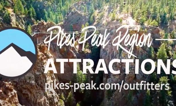 White text that says "Pikes Peak Region Attractions" with a website beneath it. Beside the text is a circular logo with a drawing of white mountains beneath blue sky. Behind the text and logo is a rope ladder stretched across and over a couple of deep valleys. The cliff tops are covered in bright green pine trees.