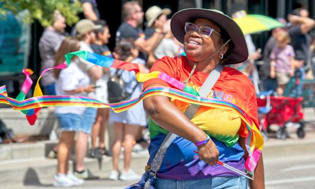 In the foreground is a person in glasses, a bucket hat, a rainbow t-shirt and jeans with a bag across their body. They are smiling peacefully with their eyes closed as they wave thin, long rainbow ribbons on sticks in front of them. In the background is a small crowd of spectators with hats, wagons and other apparel. The sun is shining.