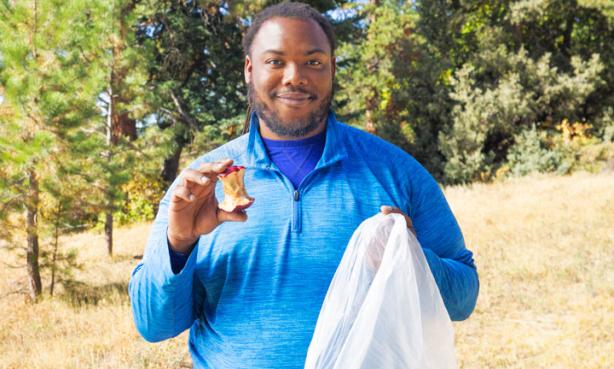 A man holds an apple core and a trash bag. He is demonstrating that you need to throw away your eaten apple instead of throwing it on the ground while hiking.