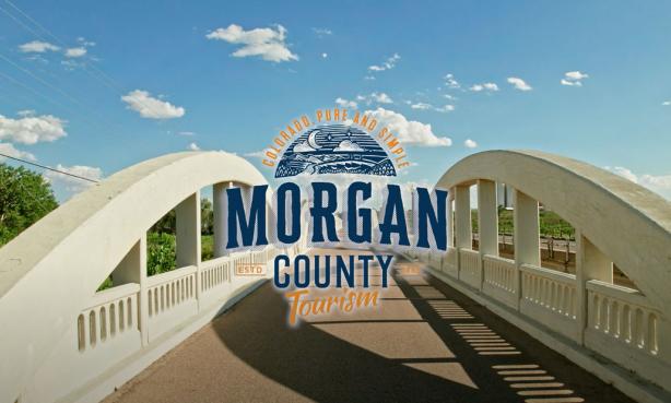 A wide shot of a white-railed bridge stretching far into the distance. Surrounding the bridge are green trees under a mostly blue sky. The words "MORGAN COUNTY Tourism" with a creative logo of mountain valleys and a moon-filled sky sit in the middle of the image.