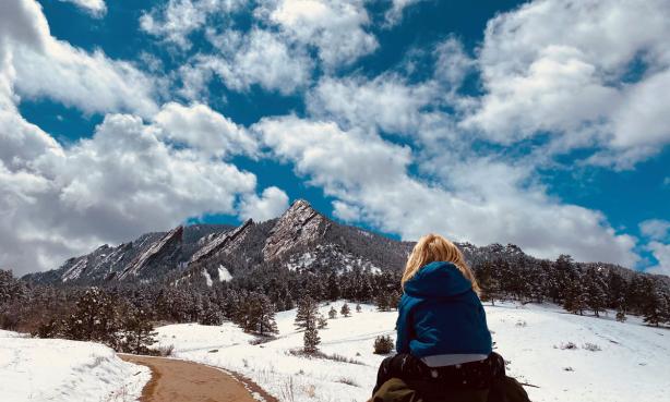 A parent carries a child on their shoulders as they hike a trail lined with snow toward the Flatirons in Boulder, Colorado. The sky is cerulean and is streaked with stark white clouds. 