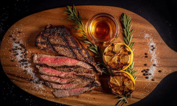 A perfectly cooked cut of steak sliced into strips on a wood platter garnished with halves of grilled lemons, fresh rosemary sprigs, sea salt, peppercorns and a shot of whiskey at The Still Whiskey Steaks in Fort Collins, Colorado