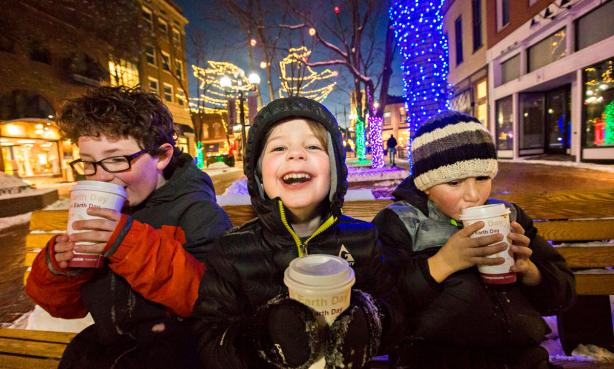 Three children sitting on a park bench on Boulder Colorado's Pearl Street. They're drinking hot cocoa and the trees in the square behind them are decked out in holiday lights.