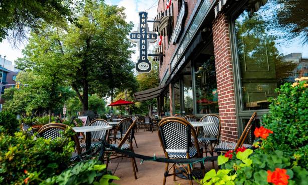 A photo of an empty patio seating outside of a brick-and-window-covered building that reads "ARMSTRONG HOTEL" on the side. There are various plants and bright-red flowers around the restaurant's patio. The restaurant is the Ace Café on College Avenue in downtown Fort Collins.
