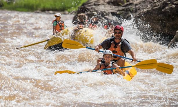 Four people smile as they paddle through the whitewater rapids of the Green River in yellow rafts. n Coloraod