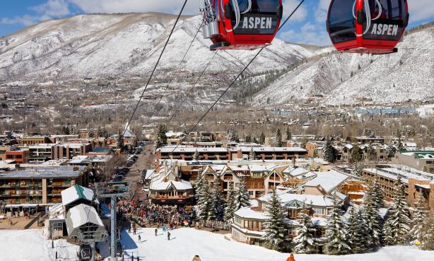 Two red and black Aspen Snowmass gondolas dangle over the ski area with hotels, mountains and a bright-blue sky in the background. Everything is covered in a layer of snow.