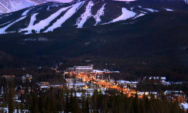 Aerial view of downtown Winter Park, with views of the snow-covered ski slopes above the town, which is lit up in the setting sun